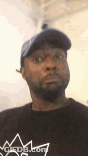 A Man With A Beard And A Hat Is Wearing A Black Shirt With A Crown On It . GIF