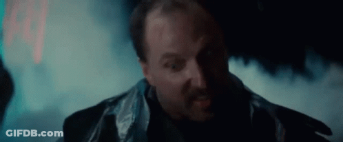 A Man With A Beard And A Leather Jacket Is Standing In A Dark Room With Smoke Coming Out Of His Mouth . GIF