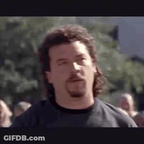 A Man With A Beard And A Mullet Is Standing In Front Of A Crowd . GIF