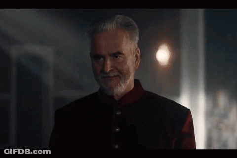 A Man With A Beard And A Red Jacket Is Standing In A Dark Room And Smiling . GIF