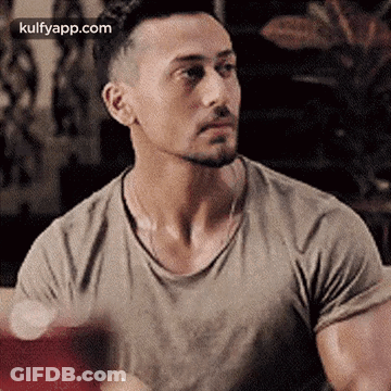 A Man With A Beard And A T-Shirt Is Sitting At A Table And Looking At The Camera . GIF