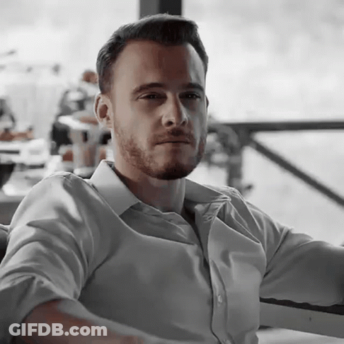 A Man With A Beard And A White Shirt Is Sitting In A Chair . GIF