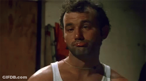 A Man With A Beard And A White Tank Top Is Making A Funny Face With His Mouth Open . GIF