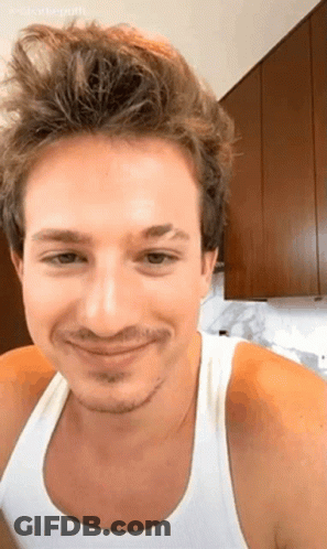 A Man With A Beard And A White Tank Top Is Smiling In A Kitchen . GIF