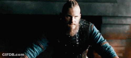 A Man With A Beard And Braided Hair Is Wearing A Leather Vest And A Blue Shirt . GIF
