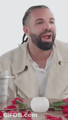 A Man With A Beard And Braids Is Sitting At A Table With Roses And A White Ball . GIF