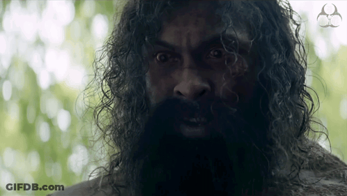 A Man With A Beard And Curly Hair Looks At The Camera With A Logo For A Company In The Background GIF