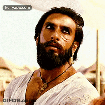A Man With A Beard And Earrings Is Wearing A White Shirt And Necklace . GIF