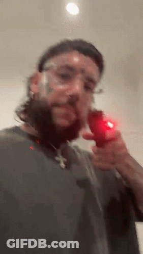 A Man With A Beard And Glasses Is Holding A Red Object In His Right Hand . GIF