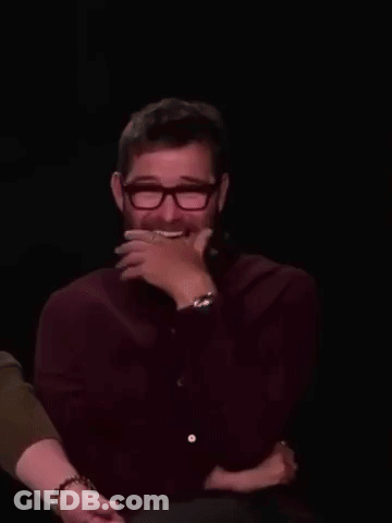 A Man With A Beard And Glasses Is Laughing With His Hand On His Face . GIF