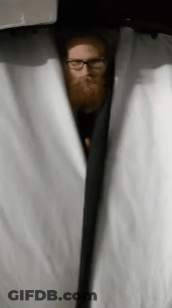 A Man With A Beard And Glasses Is Peeking Out From Behind A Curtain . GIF