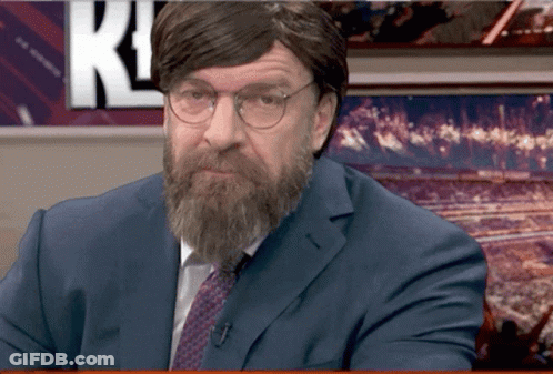 A Man With A Beard And Glasses Is Sitting In Front Of A Wall With The Letter K On It GIF