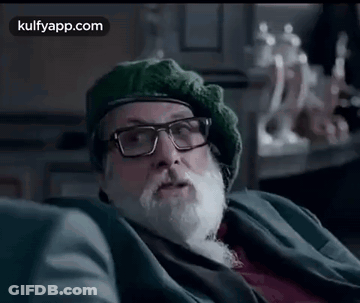 A Man With A Beard And Glasses Is Sitting On A Couch Wearing A Green Hat . GIF