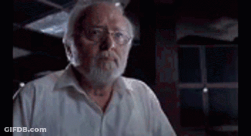 A Man With A Beard And Glasses Is Standing In Front Of A Window In A Dark Room . GIF