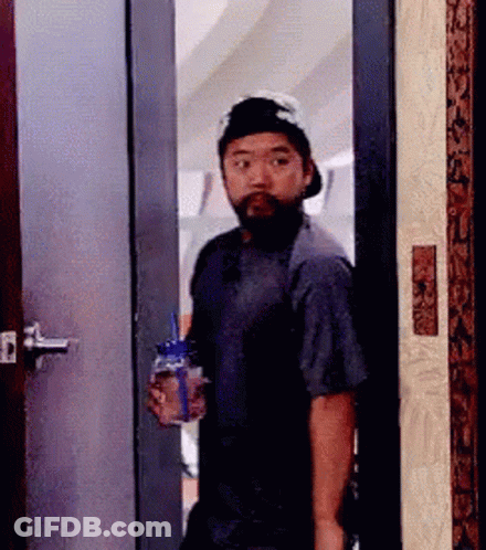 A Man With A Beard And Hat Is Standing In A Doorway Holding A Bottle Of Water . GIF