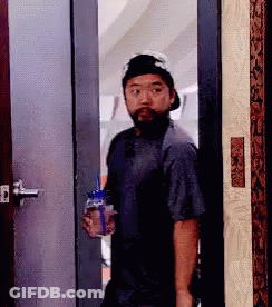 A Man With A Beard And Hat Is Standing In A Doorway Holding A Bottle Of Water . GIF