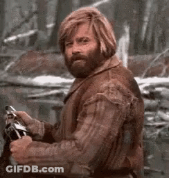 A Man With A Beard And Long Blonde Hair Is Sitting In The Woods Holding A Sword . GIF
