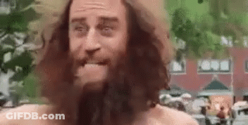 A Man With A Beard And Long Hair Is Standing In Front Of A Building And Making A Funny Face . GIF