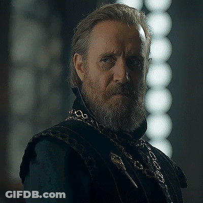 A Man With A Beard And Long Hair Is Wearing A Black Vest And A Gold Chain Around His Neck . GIF