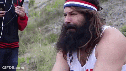 A Man With A Beard And Long Hair Is Wearing A Headband And A White Tank Top . GIF
