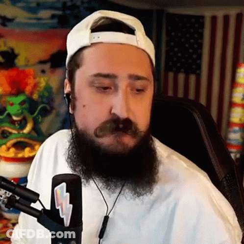 A Man With A Beard And Mustache Is Sitting In Front Of A Microphone Wearing Headphones And A White Hat . GIF