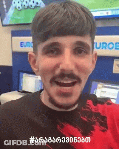 A Man With A Beard And Mustache Is Smiling In Front Of A Sign That Says Euro GIF