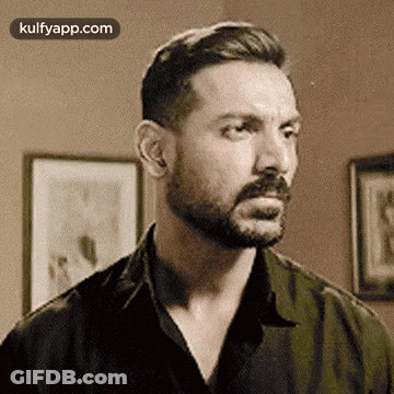 A Man With A Beard And Mustache Is Standing In Front Of A Wall With Pictures On It . GIF
