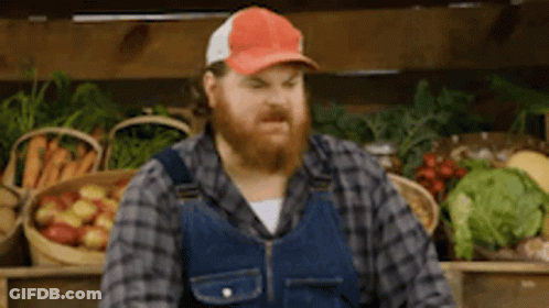 A Man With A Beard And Overalls Is Standing In Front Of A Fruit Stand . GIF
