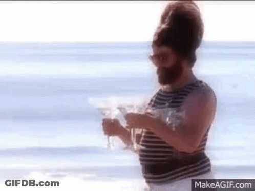 A Man With A Beard And Sunglasses Is Standing On A Beach Holding A Glass Of Water . GIF