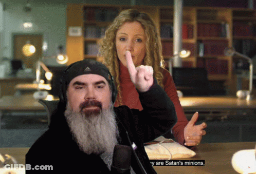 A Man With A Beard Giving The Middle Finger While A Woman Holds Her Finger To Her Mouth GIF