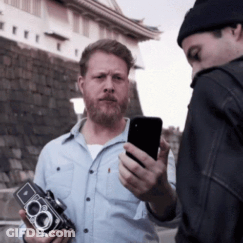 A Man With A Beard Holds A Camera While Another Man Looks At His Phone GIF