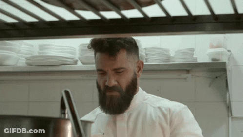 A Man With A Beard In A Kitchen With Plates On The Shelf Behind Him GIF