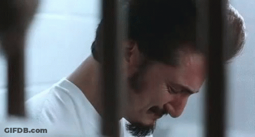 A Man With A Beard Is Behind Bars In A Jail Cell . GIF