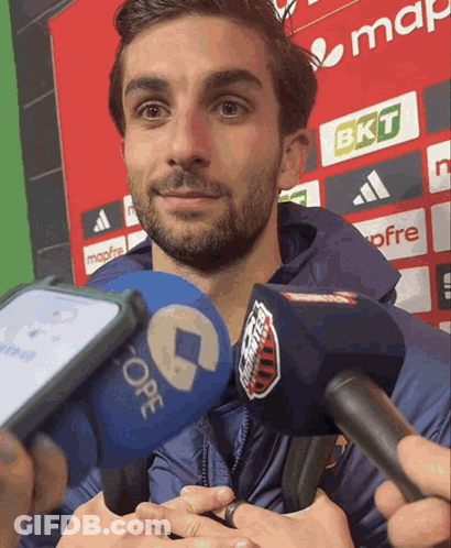A Man With A Beard Is Being Interviewed In Front Of A Sign That Says Bkt GIF