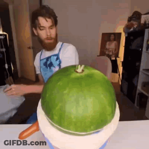 A Man With A Beard Is Carving A Watermelon With A Carrot On Top Of It . GIF