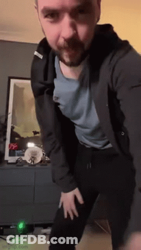 A Man With A Beard Is Dancing In A Living Room While Wearing A Black Jacket And A Blue Shirt . GIF