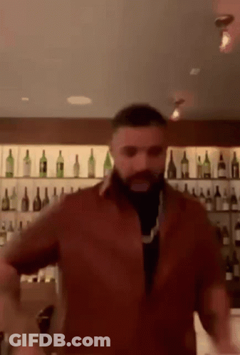 A Man With A Beard Is Dancing In Front Of A Bar Filled With Bottles Of Wine . GIF