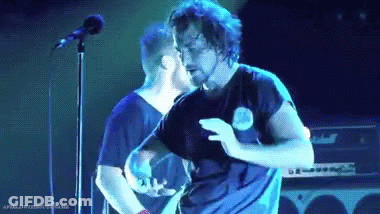 A Man With A Beard Is Dancing On A Stage In Front Of A Microphone While Another Man Stands Behind Him . GIF