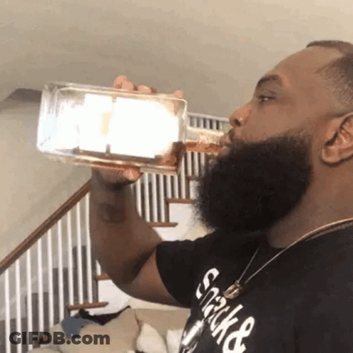 A Man With A Beard Is Drinking From A Bottle In Front Of A Staircase . GIF