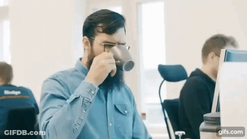 A Man With A Beard Is Drinking From A Cup In An Office . GIF