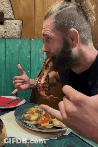 A Man With A Beard Is Eating A Large Piece Of Meat From A Plate GIF