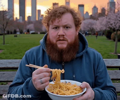 A Man With A Beard Is Eating Noodles With Chopsticks In A Park GIF