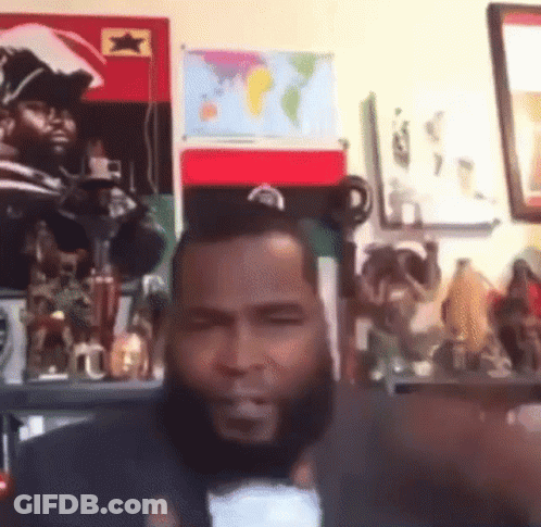 A Man With A Beard Is Giving A Thumbs Up In Front Of A Wall With A Map Of The World On It GIF