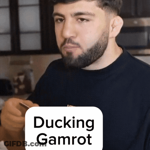 A Man With A Beard Is Holding A Cell Phone In Front Of A Sign That Reads Ducking Gamrot GIF