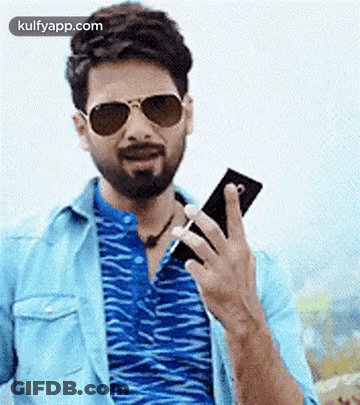 A Man With A Beard Is Holding A Cell Phone In His Hand . GIF