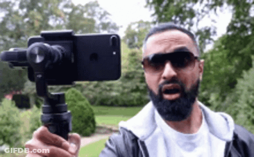 A Man With A Beard Is Holding A Cell Phone On A Tripod While Wearing Sunglasses . GIF