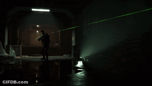 A Man With A Beard Is Holding A Gun In A Dark Room With Green Lights Coming Out Of His Face . GIF