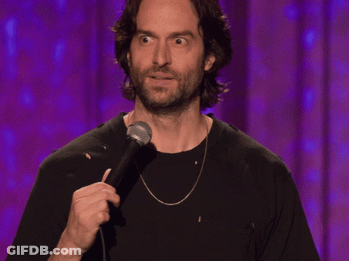 A Man With A Beard Is Holding A Microphone In Front Of A Purple Curtain GIF