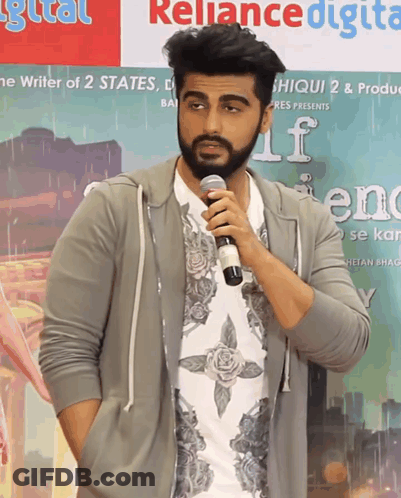 A Man With A Beard Is Holding A Microphone In Front Of A Sign That Says Reliance Digital GIF