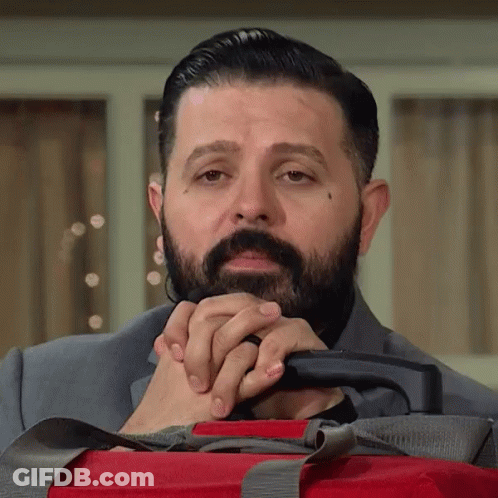 A Man With A Beard Is Holding A Red Suitcase With His Hands Folded . GIF